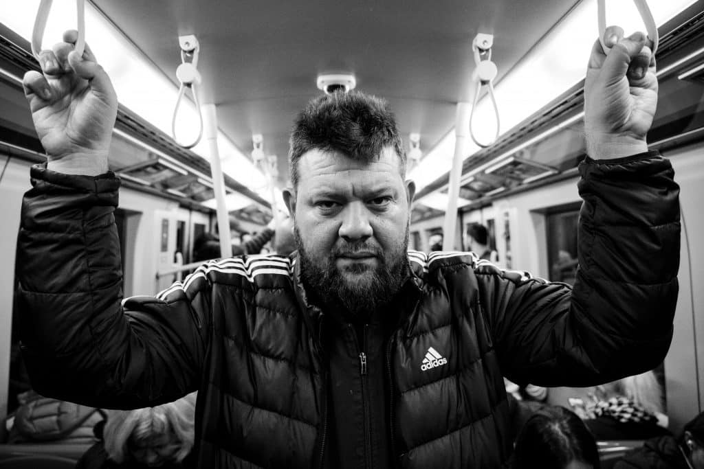 Der Künstler Kid Pex in der U-Bahn stehend. er hält sich dabei an zwi Halteschlaufen an. Das Foto ist in schwarz - Weiß gehalten.