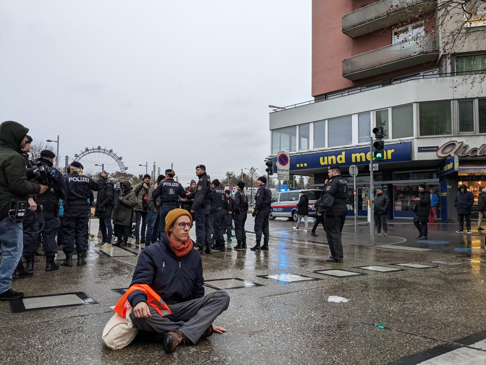 Auf dem Foto ist eine Person aus der Protestbewegung zu sehen. Die Person trägt eine blaue Jacke, eine gelbe Mütze und eine Brille. Die Person hat sich in der Nähe des Pratersterns auf dem Boden festgeklebt, um den Verkehr zu behindern.