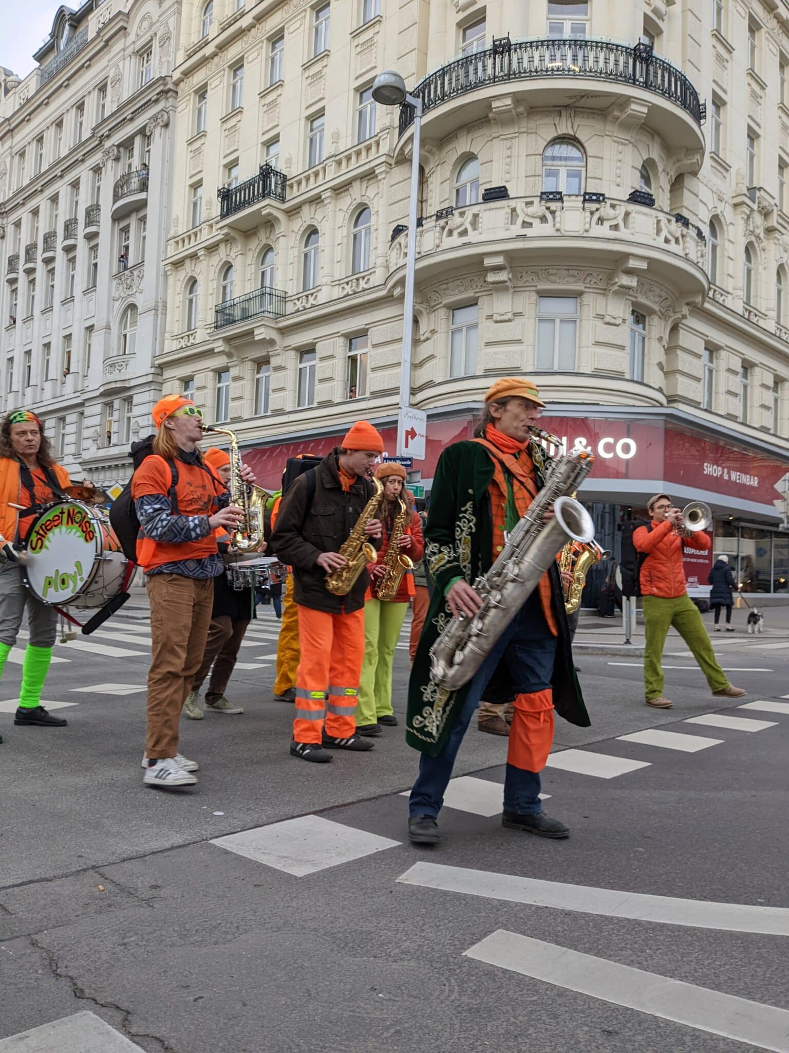 Auf dem Foto ist ein musikalischer Protest der Aktivistengruppe die "Letzte Generation Österreich" zu sehen. Dabei sind die Aktivist*innen mit Musik Instrumenten bei einem Marsch zu sehen