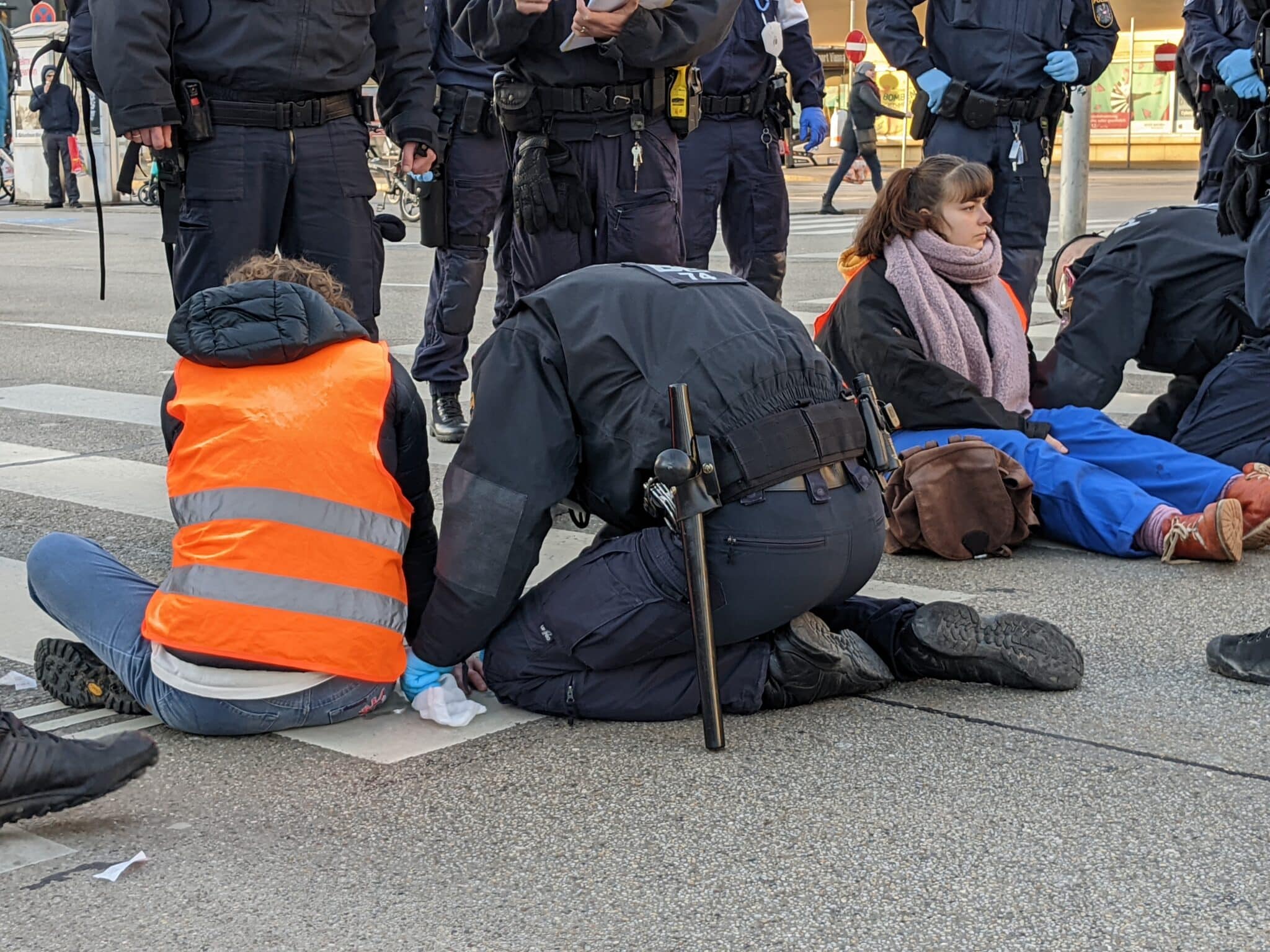 Auf dem Foto sind Polizist*innen zu sehen, wie sie Aktivist*innen von der Straße lösen. Diese hatten sich zuvor mit Sekundenkleber an die Straße geklebt, um den Verkehr zu behindern