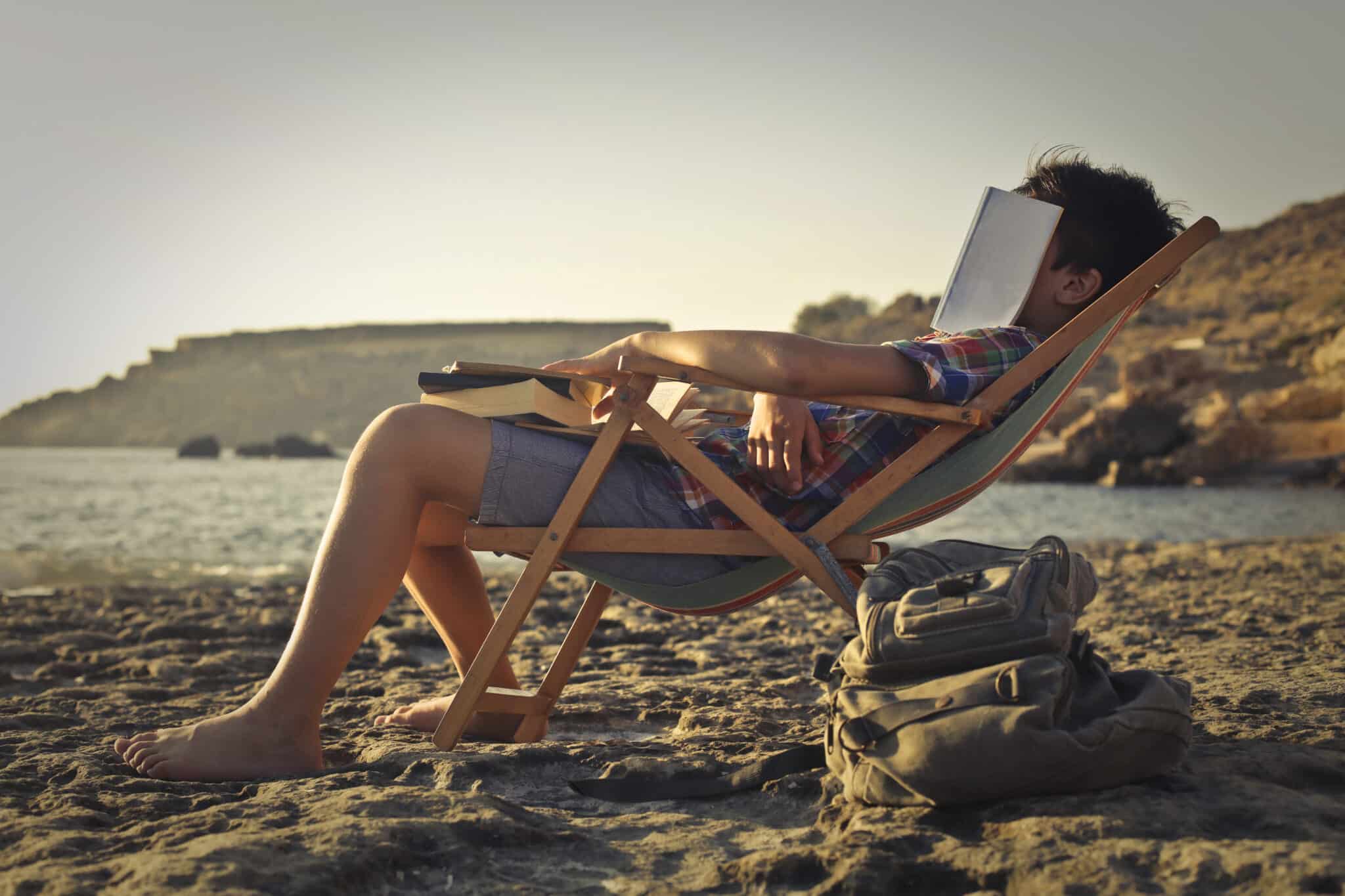 Das Foto zeigt einen Touristen am Strand. Er ist seitlich zu sehen und sitz auf einem Liegestuhl. Er scheint eingeschlafen zu sein, auf seinem Gesicht liegt ein Buch. 