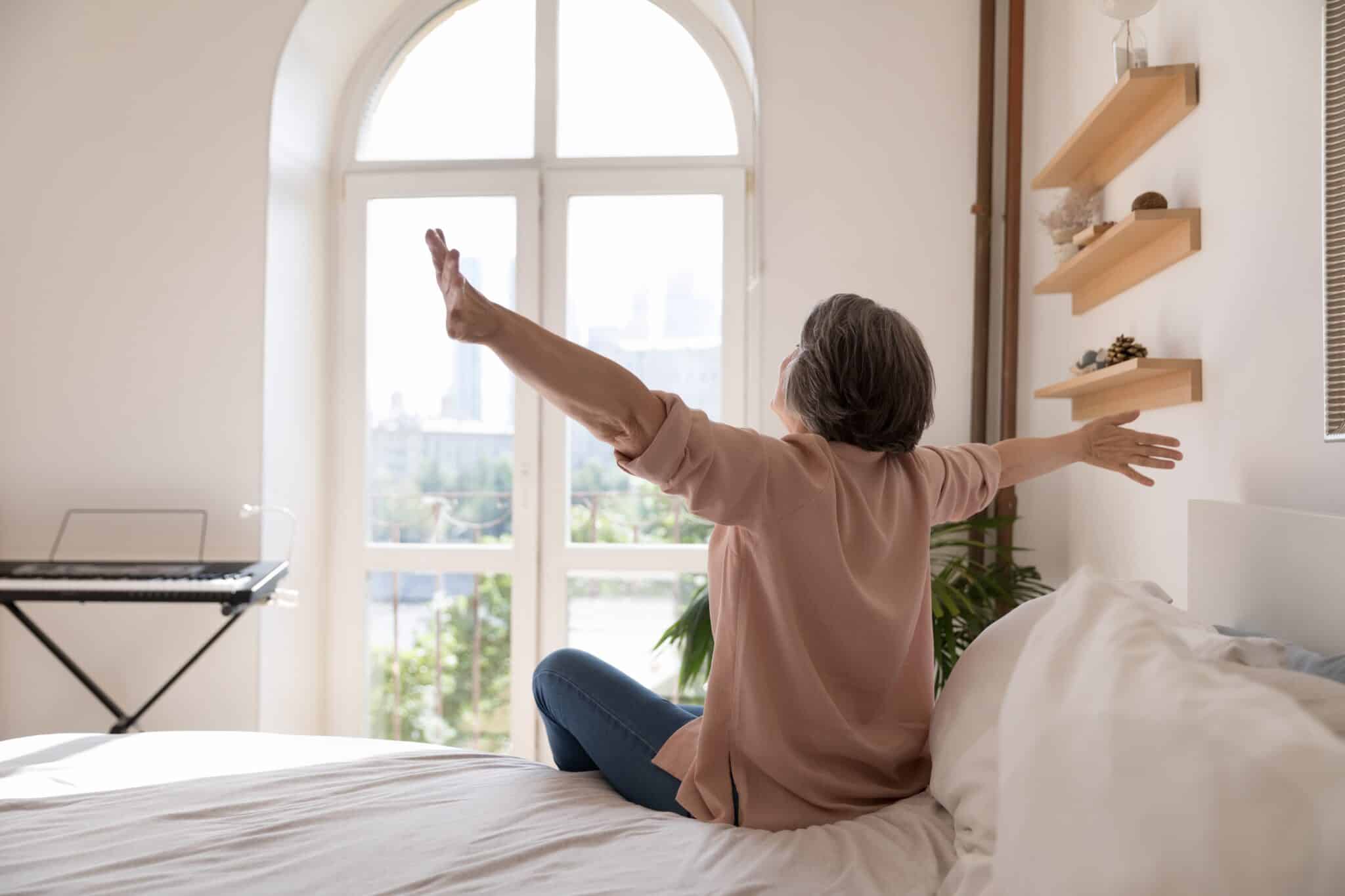 Das Foto zeigt eine Frau auf einem weißen Bett. Vor ihr ist ein Fenster und eine Keyboard zu sehen. Sie streckt dabei beide Hände nach oben.